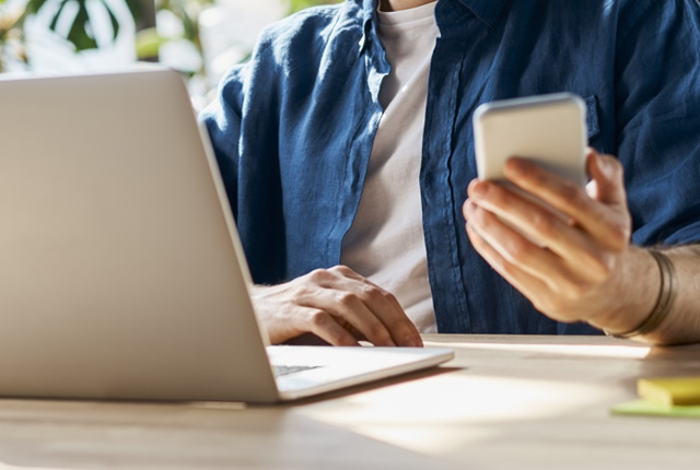 Siedzący przed laptopem mężczyzna trzyma w ręku smartfona, na którym sprawdza wymogi dotyczące usług cyfrowych związane z dyrektywą Omnibus